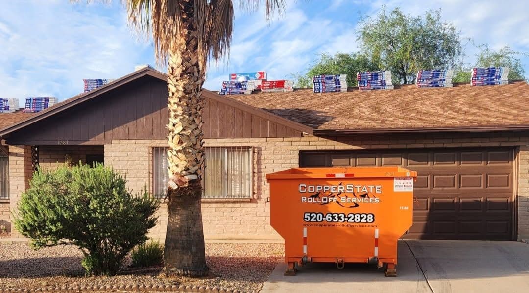 Orange dumpster in front of a residential home with roofing materials on the roof.